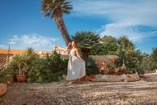 young brunette woman standing in the yard of a Spanish finca, next to a wall, wearing a cream white dress 