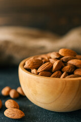 Rustic wooden bowl filled with raw almonds placed on a dark textured surface. Scattered almonds add a natural touch, complemented by a burlap fabric in the background