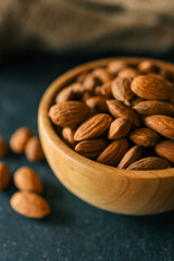 Rustic wooden bowl filled with raw almonds placed on a dark textured surface. Scattered almonds add a natural touch, complemented by a burlap fabric in the background