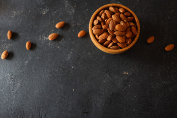 Rustic wooden bowl filled with raw almonds placed on a dark textured surface. Scattered almonds add...