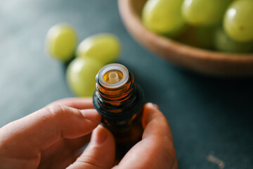 Close-up of an amber essential oil bottle being held, with green grapes in a wooden bowl in the...