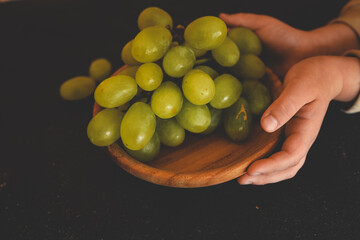 Close-up of hands holding a wooden bowl filled with fresh green grapes. Warm, rustic aesthetic highlighting the natural and wholesome appeal of the fruit