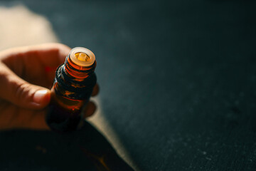 Close-up of a hand holding a small amber essential oil bottle on a dark textured surface. Minimalistic composition with warm lighting highlighting the natural tones