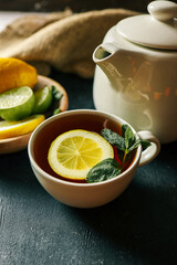 A cozy scene featuring a white ceramic teapot and a cup of freshly brewed tea garnished with a slice of lemon and fresh mint leaves. A wooden bowl with lemons and limes sits in the background
