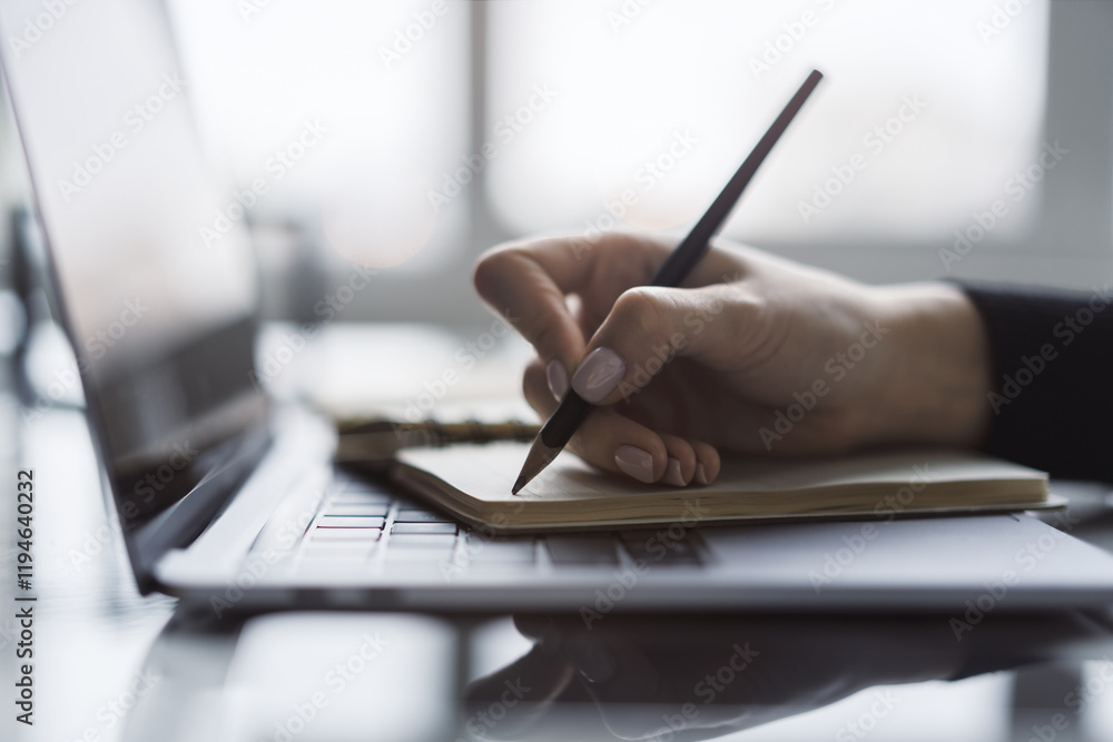 Poster The image captures a woman's hand in close view as it writes in a notepad on a chic laptop, background out of focus