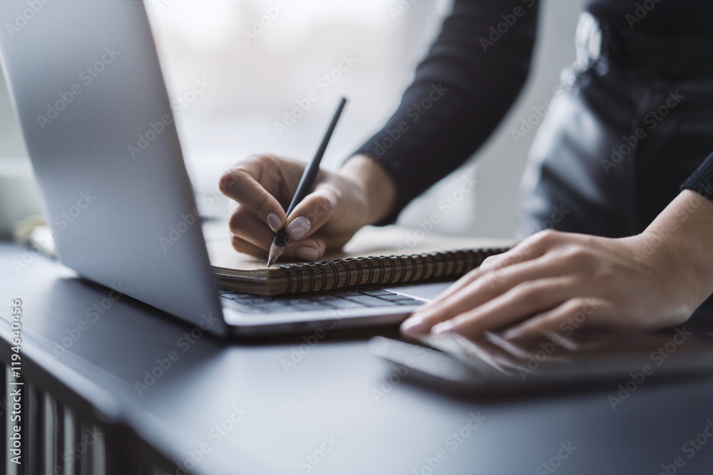 Poster close perspective of a woman's hand scribbling in a notepad on a stylish laptop, with a soft blur in