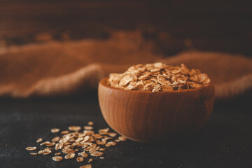Rustic wooden bowl filled with oat flakes placed on a dark textured surface with a blurred burlap...