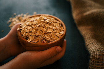 Hands holding a wooden bowl filled with rolled oats over a dark background. Some scattered oats add...