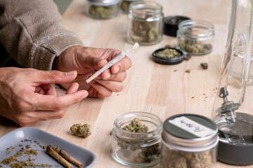 Asian man carefully rolling a cannabis joint in a cozy room, surrounded by a relaxed ambiance with cannabis equipment and accessories on the table.