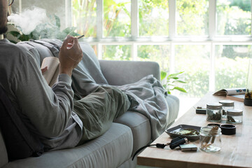 Asian man lying on a sofa, relaxing and smoking a cannabis joint in a cozy living room, with...