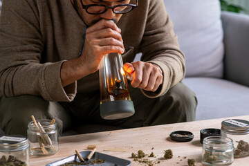 Asian man preparing and smoking a bong in a cozy living room, relaxed atmospherewith cannabis equipment and accessories on the table.
