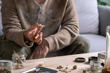Asian man smoking a cannabis joint in a cozy living room, surrounded by a relaxed ambiance with cannabis equipment and accessories on the table.