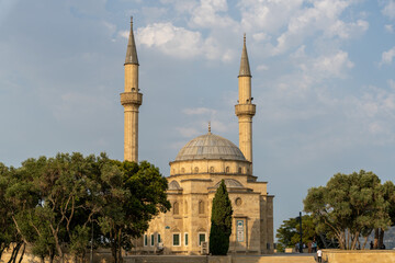 A large, old building with two tall towers and a dome. The building is surrounded by trees and he is a mosque. The scene has a peaceful and serene mood, with people walking around