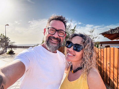 A mature couple smiling and taking a selfie with a smartphone on a sunny tropical beach during summer vacation, with clear blue ocean water, white sand, and a bright sky creating a scenic background