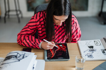 digital artist concentrates on creating intricate artwork using a stylus and tablet, seated at a well-equipped desk with sketchbooks, water, and art supplies in a modern workspace.