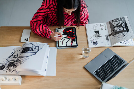 focused artist leans over a tablet, sketching an intricate design in red while surrounded by notebooks, sketches, and a modern, minimalist studio environment