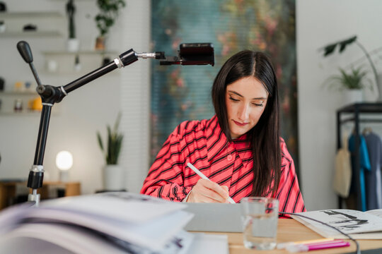 digital artist in a striped pink shirt concentrates on drawing with a stylus on a tablet, positioned under a recording smartphone in a stylish studio workspace.