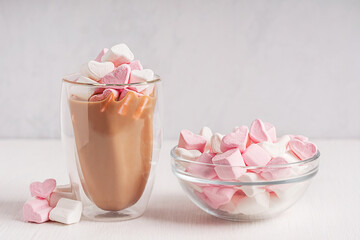 Hot chocolate, cocoa or coffee with topping of sweet organic marshmallow heart shaped dessert baked from sugar served in glass mug on white wooden table with bowl of snack for valentines day holiday