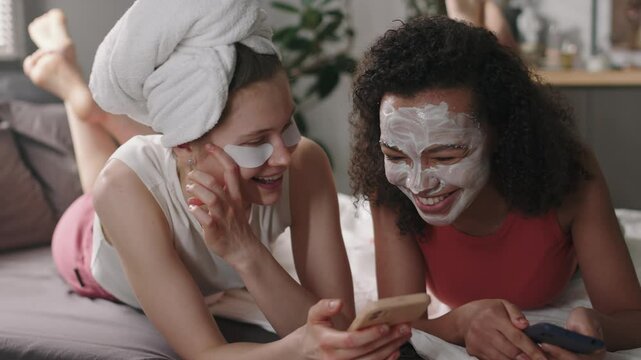Medium shot of two diverse female friends - one with towel on head and under-eye patches and other with face mask - lying in bed with phones, gossiping and laughing, discussing posts in social media