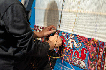 A man is sitting on the floor and working on a rug. The rug is blue and red and has a pattern of...