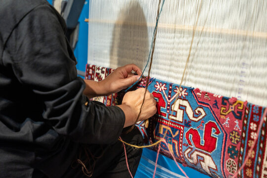 A man is sitting on the floor and working on a rug. The rug is blue and red and has a pattern of flowers. The man is using a needle and thread to stitch the rug - Powered by Adobe