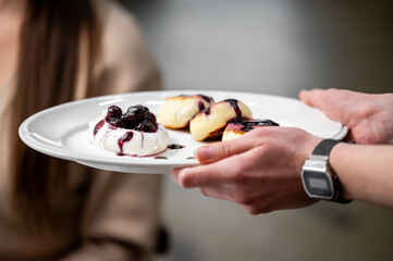 A hand holds a white plate featuring delicious pastries topped with berries and creamy yogurt. Soft focus on a person in the background adds warmth to the inviting food scene.