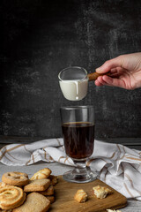 woman pouring milk from a glass milk jug into a tall glass cup with freshly brewed black coffee, Americano with milk, black background, cookies, coffee with a cookie, rustic style, freeze motion, wood
