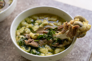 A bowl of soup with a piece of meat sticking out of it. The soup is green and white and has a lot of vegetables in it