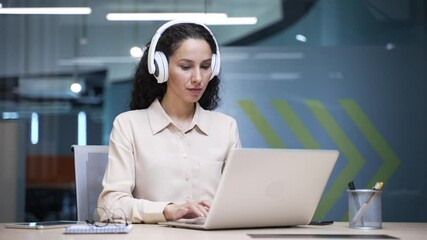 Focused businesswoman in wireless headphones typing on laptop sitting at workplace in business office. Serious thoughtful female IT specialist works on computer, thinking about solving project task