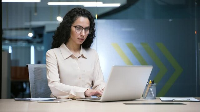Busy businesswoman working on laptop and thinking about task while sitting at workplace in modern business office. Thoughtful female employee in glasses deals with project on computer or chats online