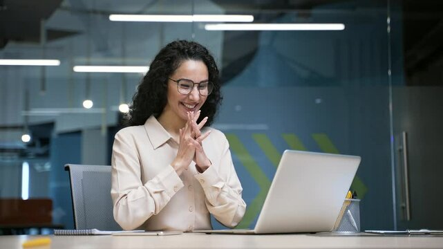 Satisfied happy businesswoman reading good news on laptop sitting at workplace in business office. Smiling glad female employee celebrates success, smiles, rejoices at positive message on computer