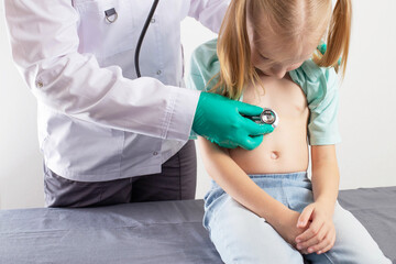 A doctor in green medical gloves listens to the bronchi with a stethoscope of a seven-year-old...