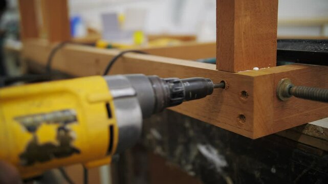 The image shows a yellow power drill being used to drive a screw into a wooden frame. The wood is clamped securely, and the background features a workshop environment with tools and materials visible.