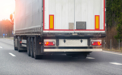 A truck with a tilt semi-trailer transports cargo along the road in the spring against the backdrop...