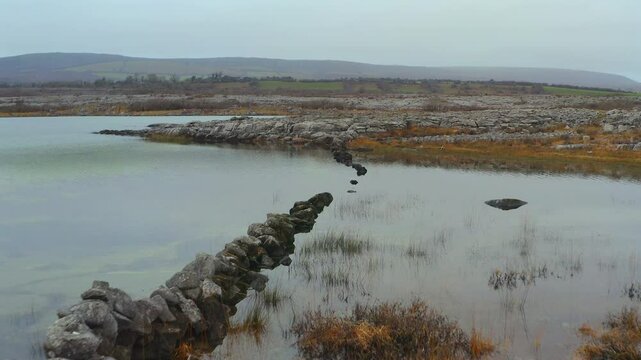 Drone glides over rugged Burren terrain, flying over a stone wall submerged in a seasonal lake