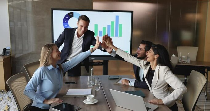 Four motivated team celebrating start-up launch, or great teamwork results, gathered together in modern conference room, stacking their hands, giving high five gesture, showing unity, shared success