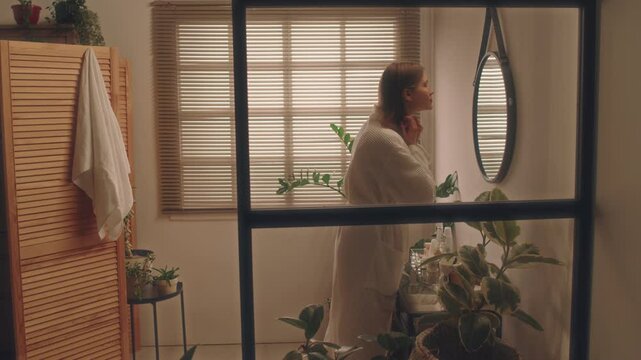 Side full shot of young Caucasian lady in white robe entering modern bathroom with biophilic design and looking at mirror, doing beauty routine in morning