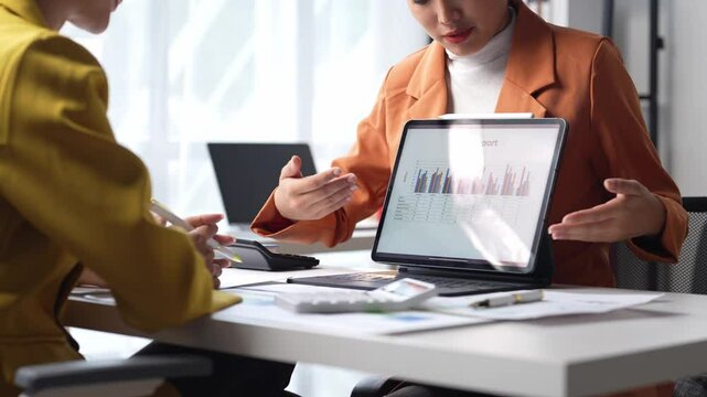 A woman in an orange jacket is holding a laptop and looking at a screen. She is in a meeting with another person, who is also looking at the laptop. The laptop screen shows a graph