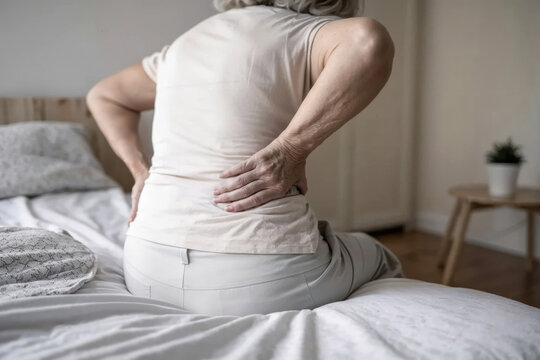A senior individual is holding their back in discomfort while seated on a bed in a bright, warm bedroom, highlighting the challenges of backache
