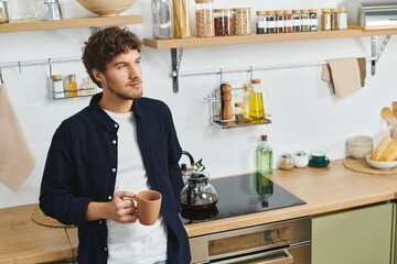 A handsome young man stands in his modern kitchen, sipping coffee and enjoying his home.