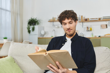 Handsome young man with curly hair relaxes while reading in his chic apartment space.