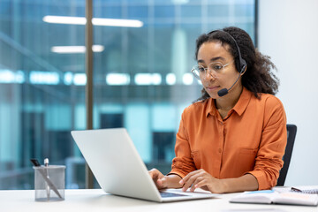 Serious focused woman working with laptop at workplace using headset phone for video call,...