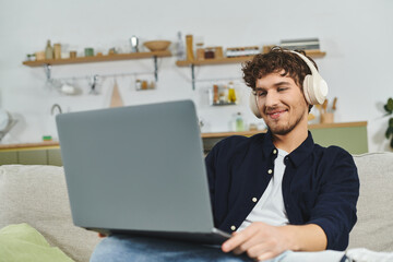 Handsome curly man relaxes on sofa with headphones, focused on his laptop in stylish living space.