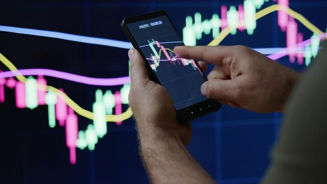 Trader or investor monitoring or looking stock market or crypto market graphics or charts on phone and PC. Man&rsquo;s hand holding phone with graphics in front of PS screen.