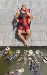 A fat man in a swimsuit sleeps by the water in which crocodiles swim