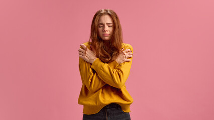 Young red-haired woman in yellow sweater suffering from fever, feeling cold and sick on pink studio...