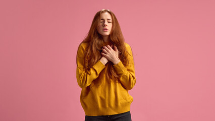 Young redhaired woman holding her chest in pain, suffering from asthma symptoms, on pink studio...