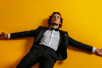 Naklejka premium A man in a suit and tie is laying on his back on a yellow wall. The man is wearing a black suit and a white shirt. He is wearing a black bow tie. The man is relaxed and comfortable in his attire