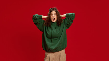 Shocked woman, with curly hair, dressed casual attire, posing holding hands on head and with open mouth looking at camera against red background. Concept of human emotions, self-expression, fashion.