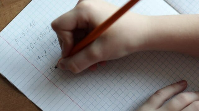 a child writes with a pencil and solves math examples in a checkered notebook. primary school education. close-up